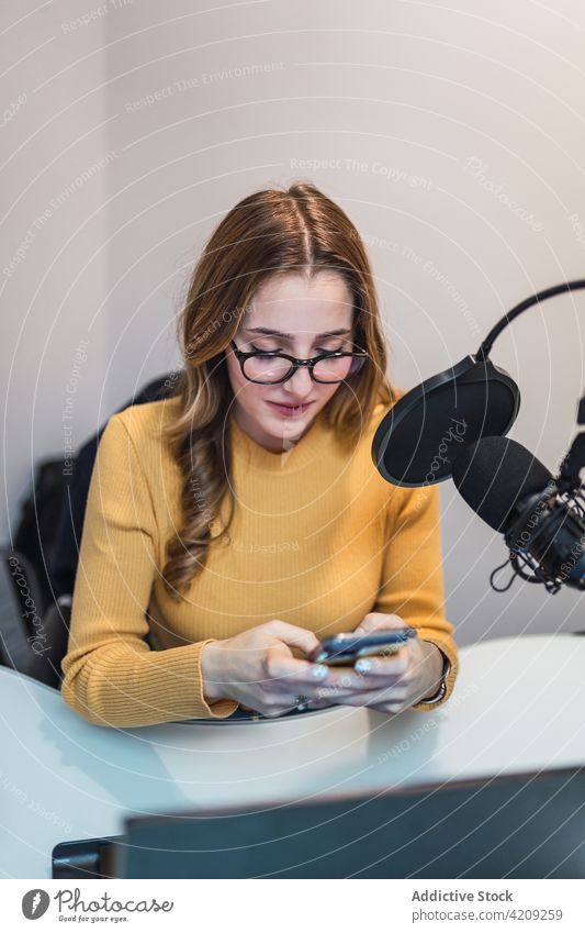 Zufriedene Frau, die im Radiosender ihr Smartphone durchsucht Textnachricht Arbeit Ausstrahlung Atelier Laptop Mikrofon Browsen Gerät Surfen modern SMS