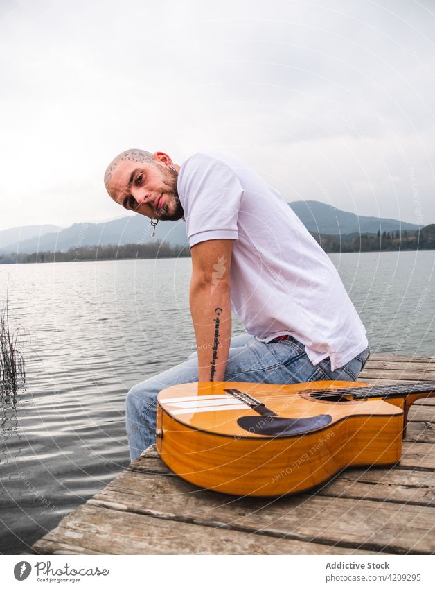 Moderner Mann mit Gitarre auf einem Steg am See Pier Fluss Natur Musiker Berge u. Gebirge hölzern wolkig grau Himmel Gitarrenspieler tagsüber friedlich Wasser