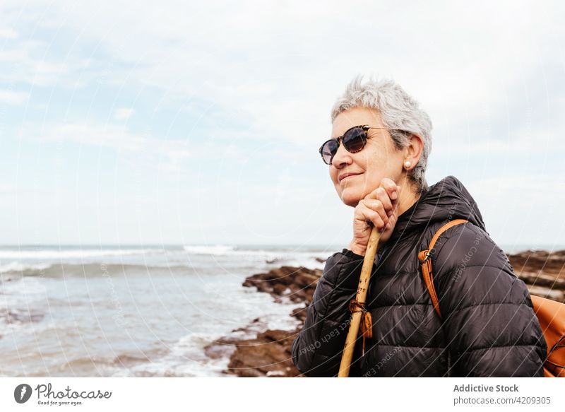 Zufriedener älterer Wanderer bewundert das Meer unter bewölktem Himmel bewundern Wanderung Lächeln genießen Natur Meereslandschaft Frau Porträt nachdenken