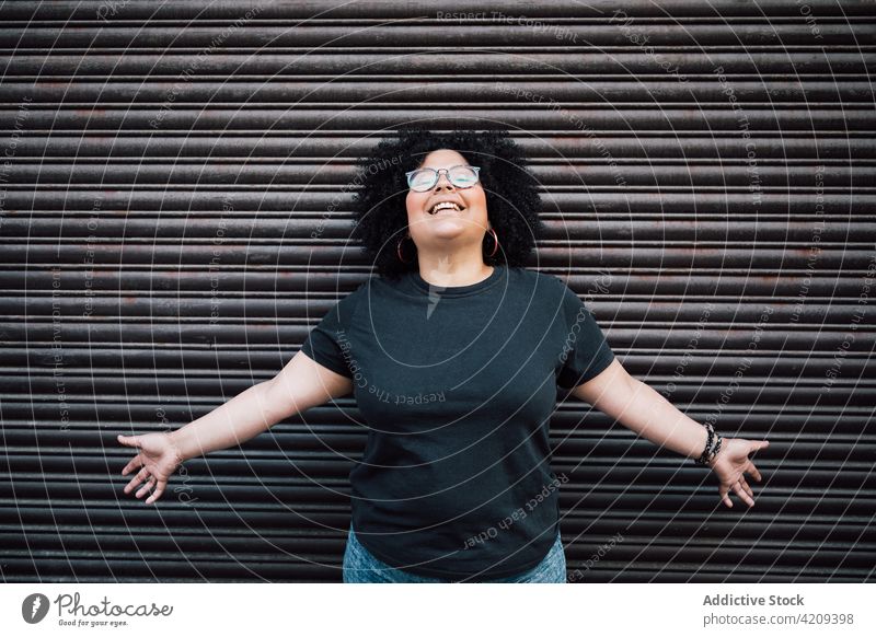 Glückliche, mollige Frau mit offenen Armen an einer gerippten Wand Lachen herzlich freundlich Brille Afro-Look charmant Porträt Übergewicht genießen Lächeln