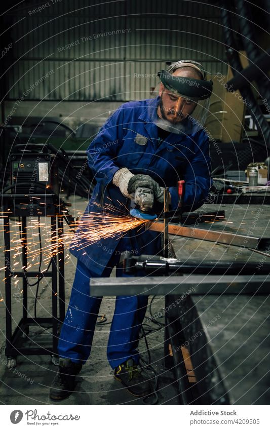 Ein Mann bearbeitet ein Metallteil in einer Werkstatt Schweißer Winkelschleifer Grinden Funken Arbeiter Konstruktion professionell Arbeitsbekleidung Mundschutz