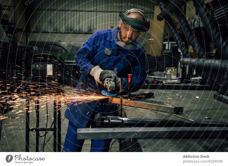 Ein Mann bearbeitet ein Metallteil in einer Werkstatt Schweißer Winkelschleifer Grinden Funken Arbeiter Konstruktion professionell Arbeitsbekleidung Mundschutz
