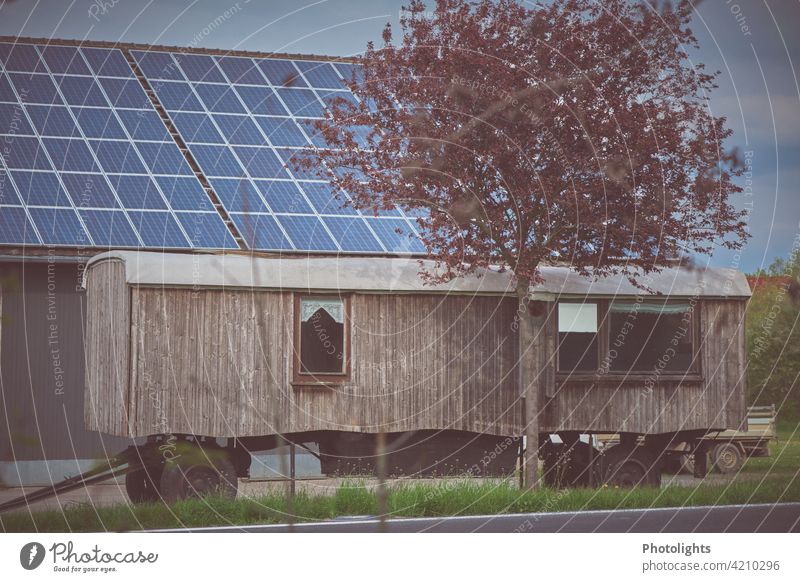 Holzwohnwagen steht vor einer Scheune mit Solardach. Blühender Baum im Vordergrund. Bauwagen Anhänger Wohnwagen Vorhang Räder Anhängerkupplung blühend Blüten