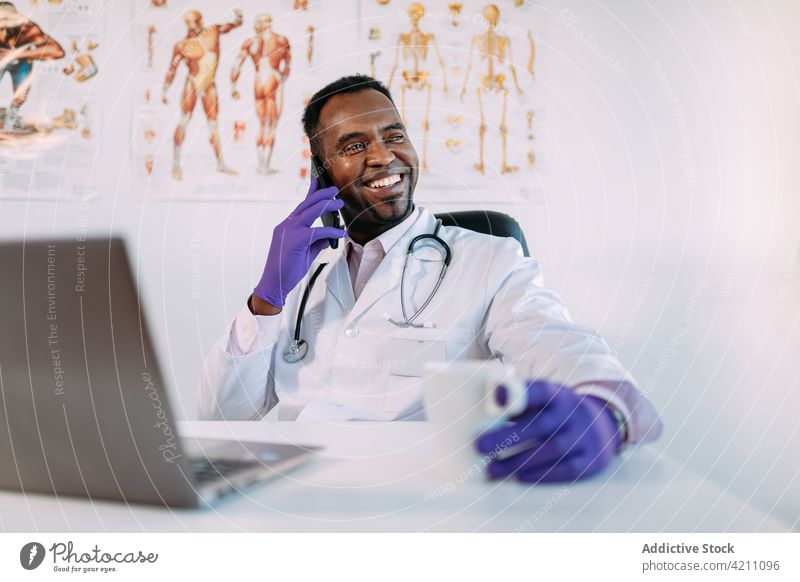 Glücklicher schwarzer männlicher Arzt, der mit seinem Smartphone spricht und Kaffee im Krankenhaus trinkt Mann Telefonanruf Lächeln Kaffeepause arzt heiter