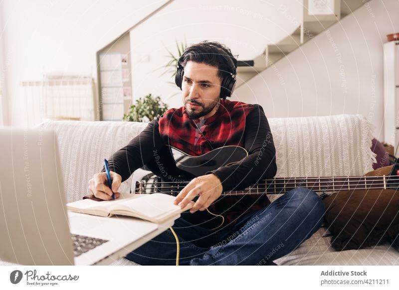 Bassist mit Gitarre schreibt in Notizblock gegen Laptop im Haus schreiben komponieren kreieren Kopfhörer zuhören Mann benutzend Apparatur Gerät
