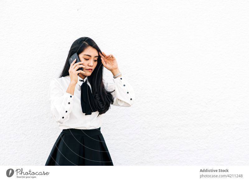 Asiatische Frau, die mit ihrem Smartphone gegen eine weiße Wand spricht Schüler Telefonanruf benutzend Teenager reden Internet Mitteilung elegant nachdenklich