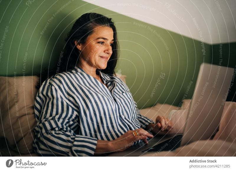 Selbstbewusste reife hispanische Dame, die im Bett aus der Ferne an einem Netbook arbeitet Frau Arbeit Laptop freiberuflich abgelegen Lächeln Inhalt online