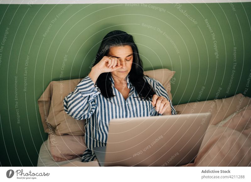 Müde ethnische Frau reibt sich die Augen bei der Online-Arbeit am Laptop auf dem Bett erschöpft abgelegen freiberuflich Auge reiben müde Überarbeitung Job