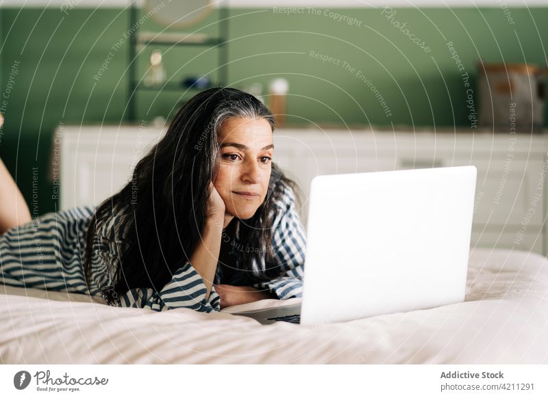 Ethnische Frau liegt auf dem Bett und arbeitet aus der Ferne an einem Netbook Laptop Lügen freiberuflich Arbeit abgelegen Internet Anschluss digital reif