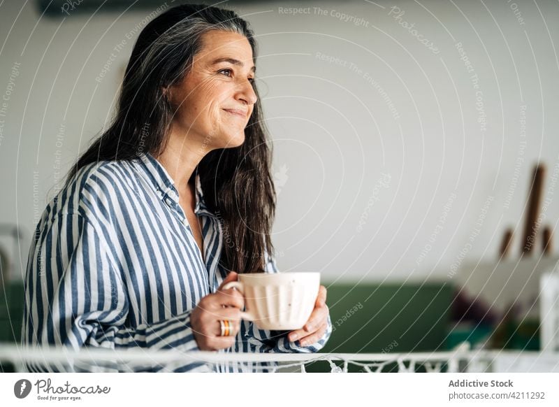Positive reife Frau mit Kaffee im Zimmer Tasse Raum heimisch trinken Freizeit Erholung Malerei Lebensmitte Staffelei Zeichnung Heißgetränk Getränk Lächeln