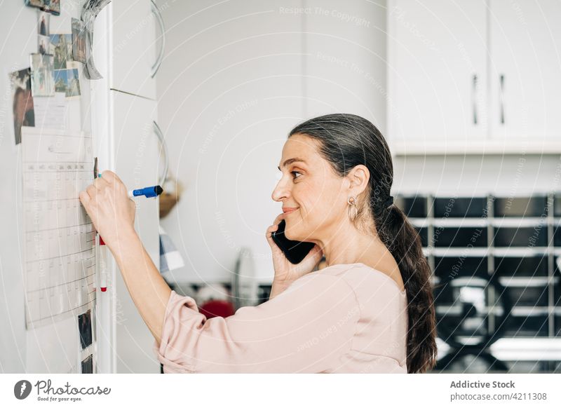 Zufriedene reife Frau, die telefoniert und sich Notizen auf Papier am Kühlschrank macht Kalender zur Kenntnis nehmen Smartphone Küche reden schreiben Gespräch