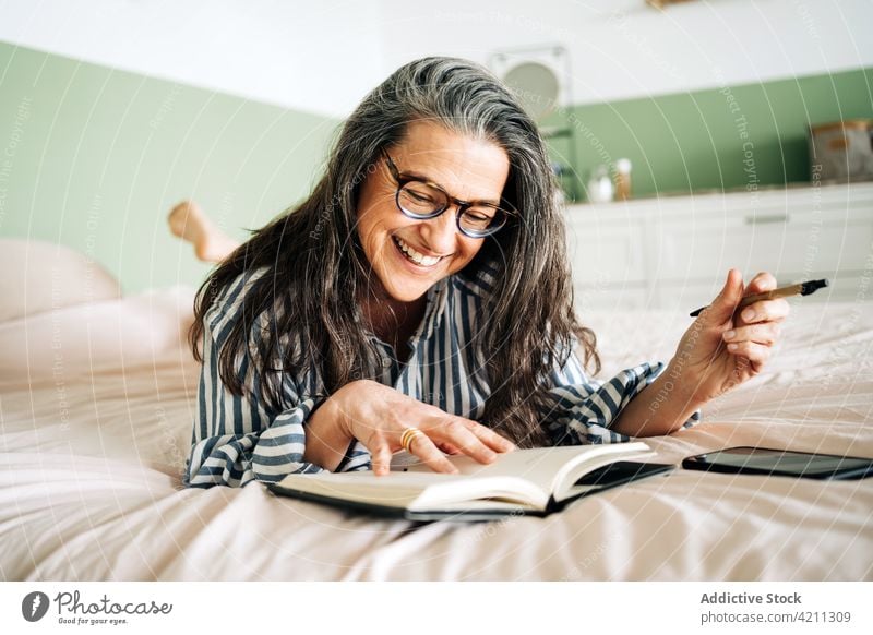 Reife glückliche Frau überprüft Planer auf dem Bett heiter Zeitplan Notebook Smartphone Lächeln lesen reif Streifen prüfen Hemd Lebensmitte trendy positiv