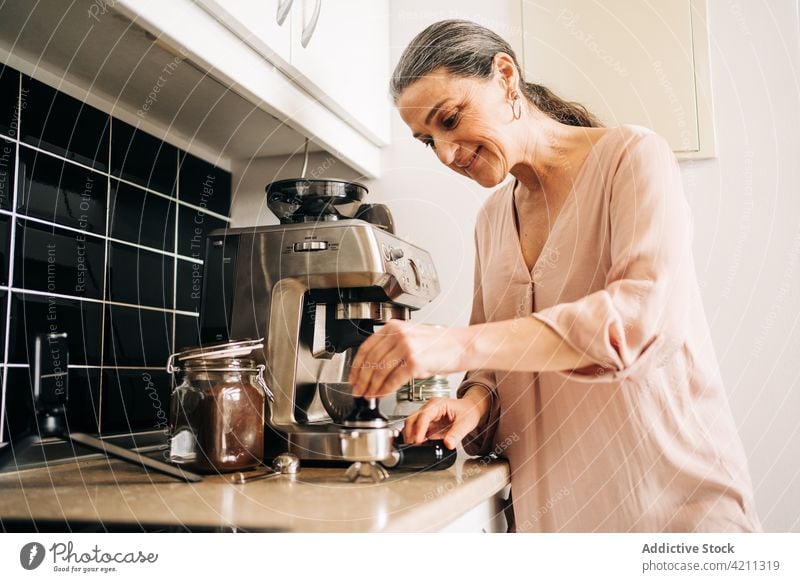 Konzentrierte reife Frau, die ein Getränk mit einer Kaffeemaschine zubereitet vorbereiten Smartphone Tasse Küche Gerät Küchengeräte heimisch trinken Schrank