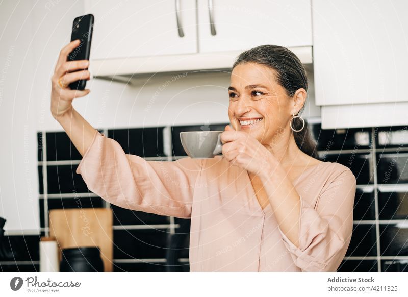 Fröhliche reife Frau mit Kaffee, die ein Selfie in der Küche macht Selbstportrait Smartphone Tasse Fotografie trinken Schrank heimisch Funktelefon Lebensmitte