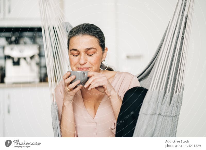 Fröhliche reife Frau trinkt Kaffee und sitzt in der Hängematte Küche Tasse Raum trinken heimisch Freizeit Erholung Lebensmitte Augen geschlossen Heißgetränk