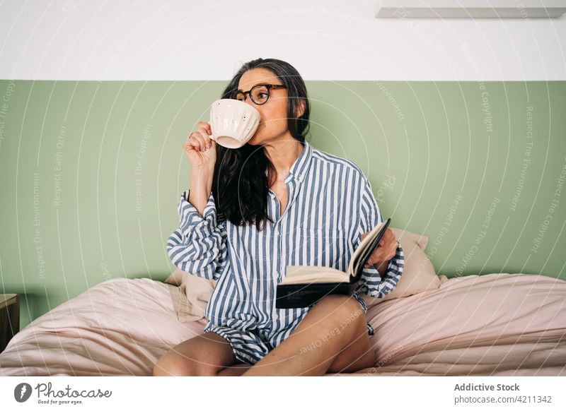 Frau mit Planer trinkt Kaffee auf dem Bett Tasse trinken Notebook Brille friedlich Freizeit Streifen Hemd heimwärts Schlafzimmer weich ruhig klug Komfort Dame
