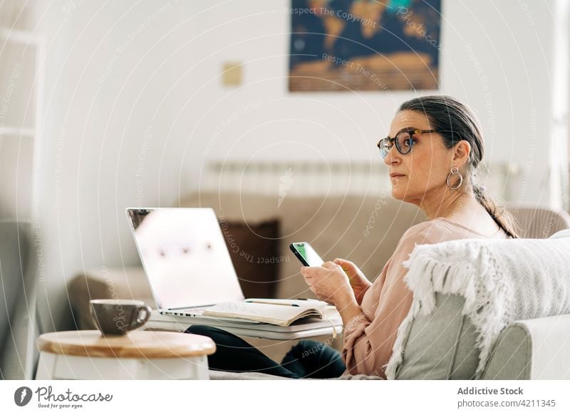 Seriöse reife Frau am Handy und am Computer arbeitend Smartphone Kommunizieren Laptop Internet Browsen Lebensmitte Nachricht online Sofa Raum Funktelefon