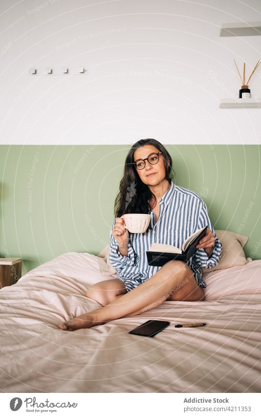 Frau mit Planer trinkt Kaffee auf dem Bett Tasse trinken Notebook Brille friedlich Freizeit Streifen Hemd heimwärts Schlafzimmer weich ruhig klug Komfort Dame