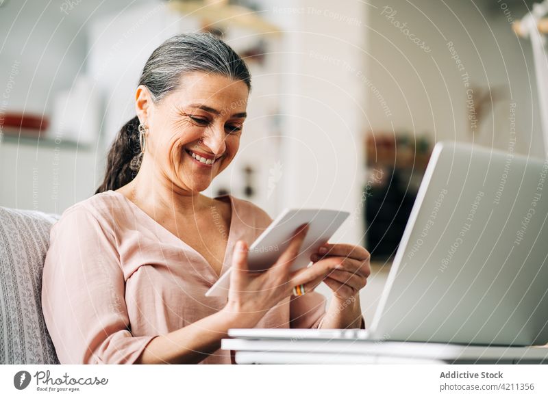Lächelnde reife Frau betrachtet ein Foto in der Nähe eines Laptops bewundern untersuchen Lebensmitte Sofa Raum heimwärts Netbook Küche Apparatur Gerät genießen