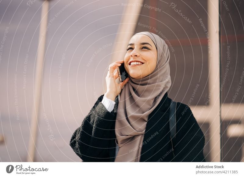 Fröhliche muslimische Frau, die in der Stadt mit ihrem Smartphone spricht sprechen reden Großstadt Hijab Lächeln Gespräch Telefonanruf Straße ethnisch Mobile