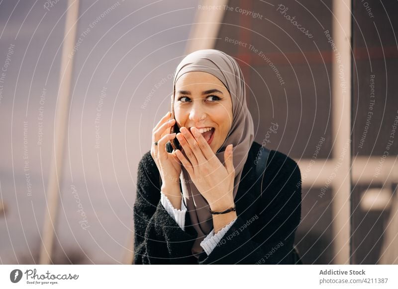 Fröhliche muslimische Frau, die in der Stadt mit ihrem Smartphone spricht sprechen reden Großstadt Hijab Lächeln Gespräch Telefonanruf Straße ethnisch Mobile