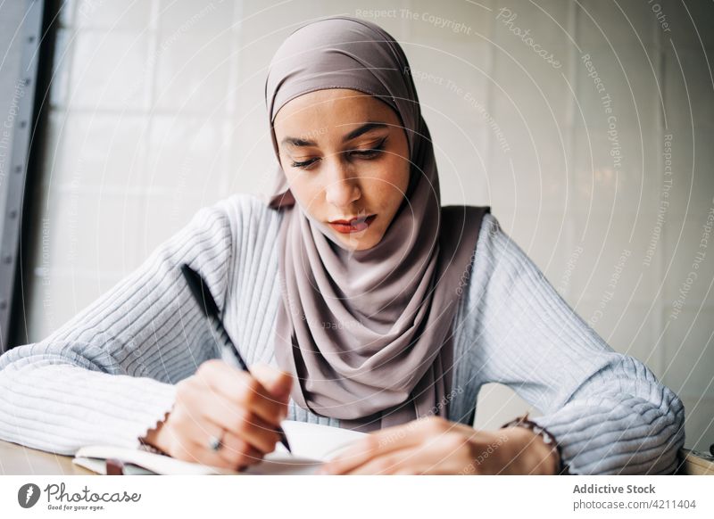 Ernste muslimische Frau, die in einem Café Notizen in ein Notizbuch schreibt zur Kenntnis nehmen Notebook freiberuflich Arbeit abgelegen schreiben Notizblock