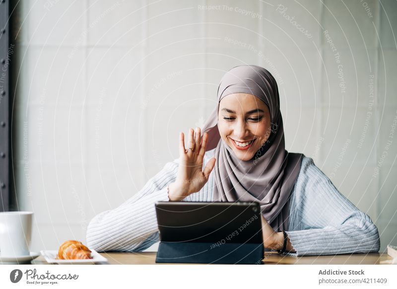 Entzückte ethnische Frau mit Videoanruf auf dem Tablet Video-Chat Wellenhand Gruß Hallo Tablette reden Hi Hijab muslimisch Tisch Café Inhalt heiter
