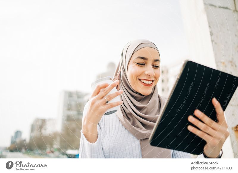 Lächelnde ethnische Frau im Hidschab, die in der Stadt ein Tablet benutzt Browsen Tablette Großstadt Imbissbude trinken benutzend Kopftuch Straße muslimisch