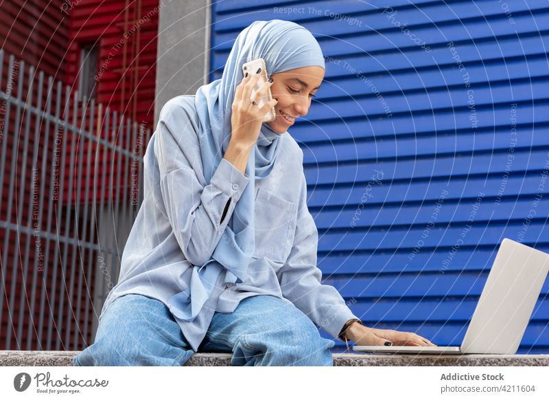 Fröhliche ethnische Frau mit Laptop, die in der Stadt mit ihrem Smartphone spricht reden Multimedia heiter Zeitvertreib urban Porträt benutzend Apparatur Gerät