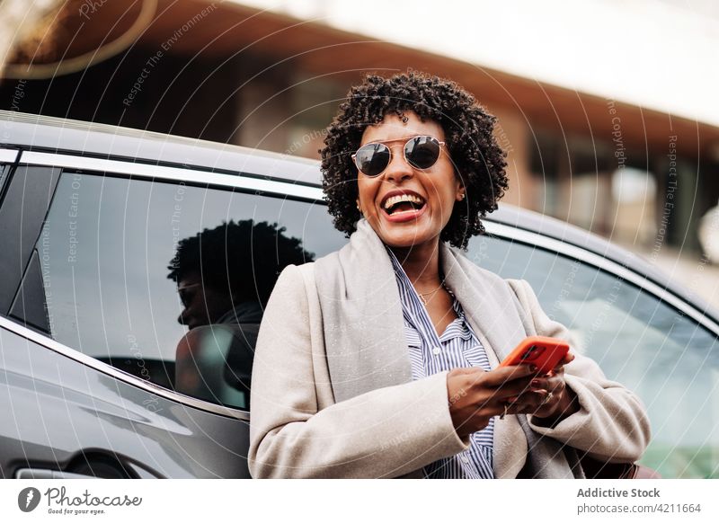 Vergnügte schwarze Frau, die in der Nähe ihres Autos auf ihrem Smartphone surft benutzend Lachen Glück Stil Freude sorgenfrei PKW Gerät Browsen heiter