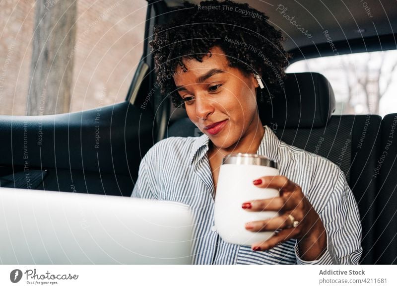 Junge schwarze Frau mit Laptop im Auto benutzend Kaffee Morgen Arbeitsweg Passagier Taxi Tasse heiter Inhalt Rücksitz tws echte drahtlose Ohrstöpsel PKW