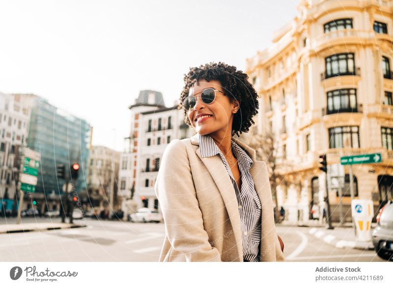 Fröhliche schwarze Frau mit trendiger Sonnenbrille lächelt auf der Straße heiter trendy Stadt Lächeln positiv Glück Mantel urban Mode Outfit Großstadt jung froh