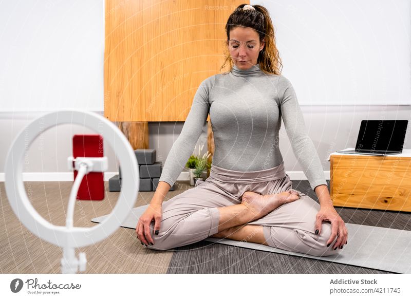 Meditierende Frau in der Yogastellung Padmasana padmasana Lotus-Pose meditieren vlog Augen geschlossen Smartphone Stativ Blogger üben jung Sportkleidung