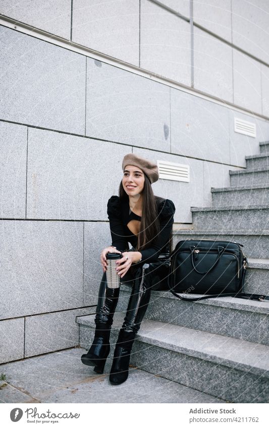 Lächelnde Geschäftsfrau auf einer Treppe sitzend mit einem Getränk zum Mitnehmen Großstadt Imbissbude trinken Straße genießen Stil trendy Frau Baskenmütze