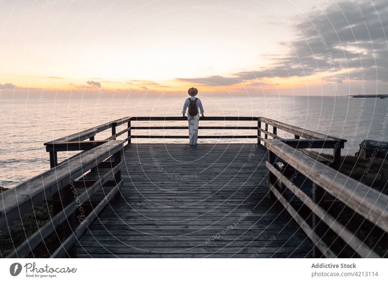 Rückenansicht Mann mit Rucksack und Hut, der auf einem Gehweg steht und den Sonnenaufgang am Meer betrachtet MEER Laufsteg Wasser Natur Freizeit Blick