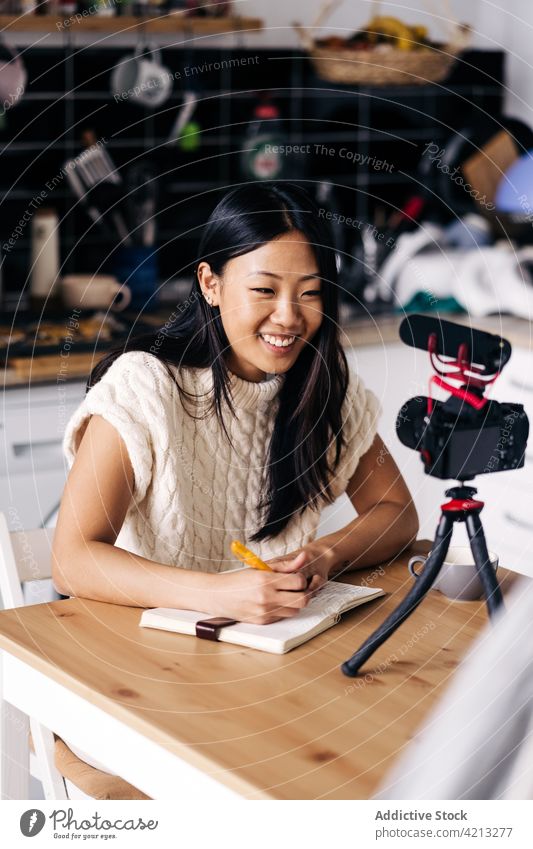 Glückliche asiatische Bloggerin mit Tagebuch und Fotokamera zu Hause Notebook Fotoapparat Lächeln vlog vorbereiten Frau Tisch heiter Küche Vlogger Video