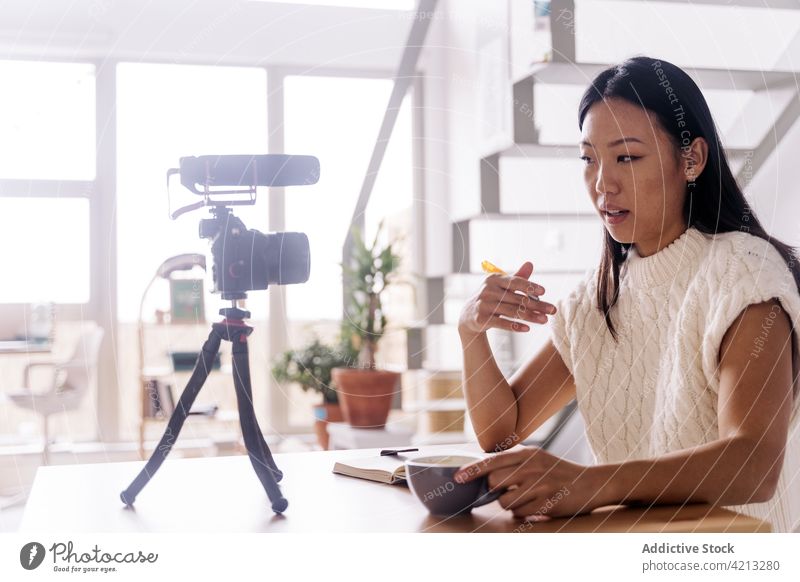 Asiatischer Blogger mit Tagebuch und Fotokamera zu Hause Notebook Fotoapparat vlog achtsam vorbereiten ernst Frau Tisch Küche Vlogger blanko Papier Fokus