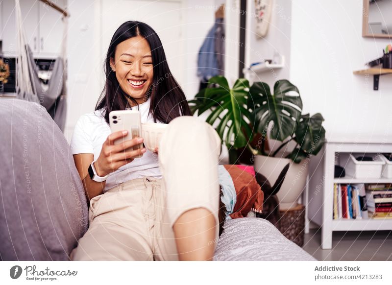 Lächelnde asiatische Frau, die zu Hause auf dem Sofa mit ihrem Smartphone chattet Textnachrichten Internet online heiter freie Zeit Porträt benutzend Apparatur