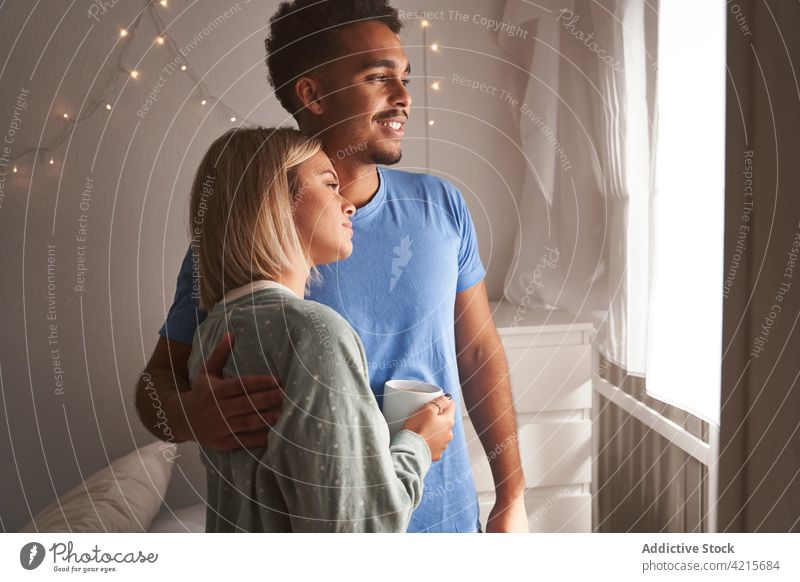 Sanftes Paar, das sich am Morgen zu Hause umarmt Umarmung Zusammensein Schlafzimmer Fenster Angebot Pyjama Umarmen heimwärts multiethnisch rassenübergreifend