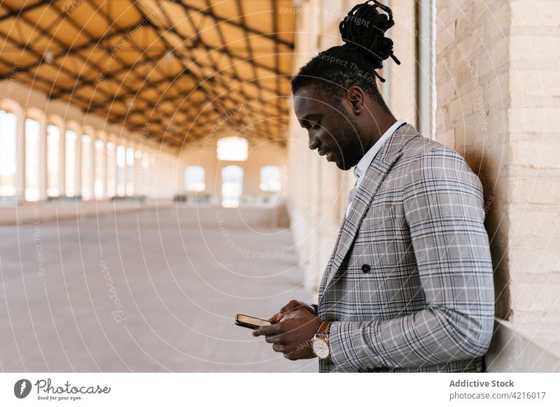 Stilvoller schwarzer Geschäftsmann, der mit seinem Smartphone gegen eine Hauswand chattet plaudernd stylisch Individualität Rastalocken Internet Gebäude Porträt