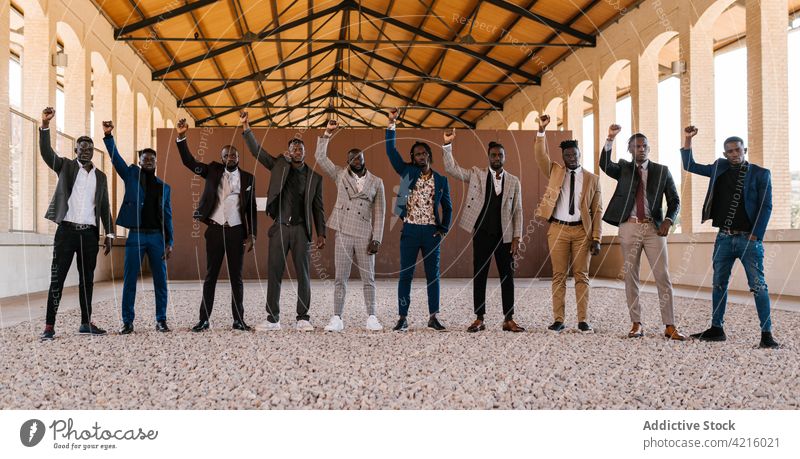 Schwarze Geschäftsleute in Anzügen mit erhobenen Armen mit der Faust nach oben Männer Stil Mode Anzug auflehnen Revolution selbstsicher Porträt Gebäude