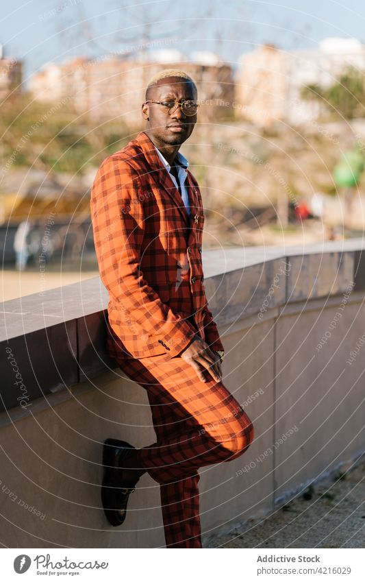 Stilvoller ethnischer Mann im Anzug vor städtischem Zaun Mode Individualität kariert maskulin Bein angehoben Stadt Tartan Porträt Brille cool gut gekleidet