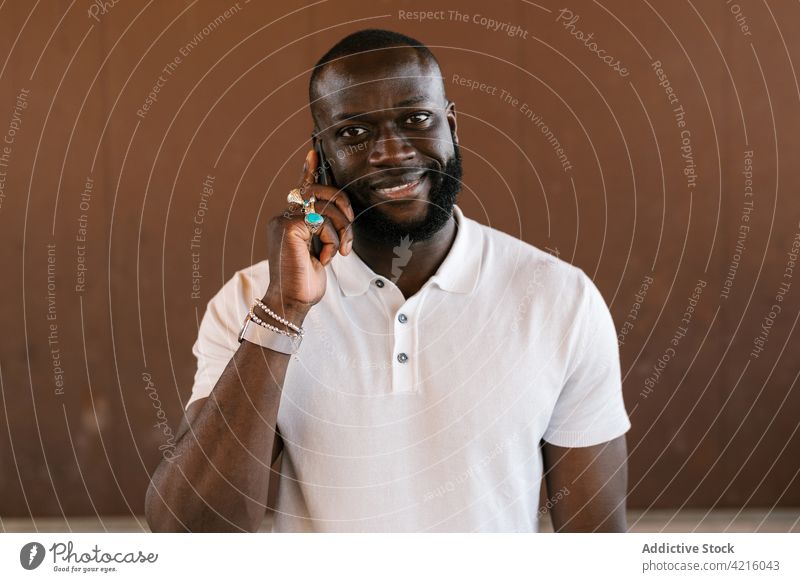 Lächelnder schwarzer Mann in Ringen, der mit einem Smartphone spricht sprechen senden heiter Stil Telefonanruf Accessoire reden benutzend Apparatur Gerät