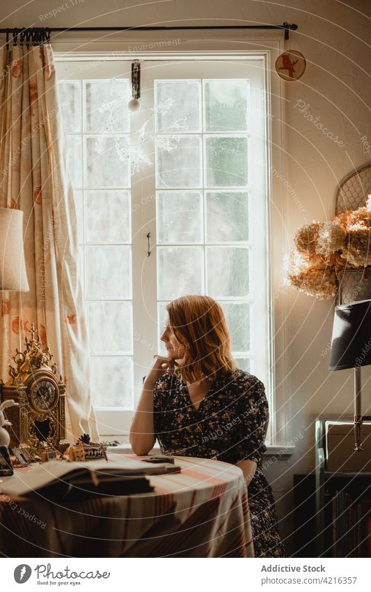 Verträumte Frau am Tisch mit antiker Uhr zu Hause verträumt Dekor Antiquität Buch Fenster idyllisch Harmonie genießen Achtsamkeit retro Stil Pflanze glänzend