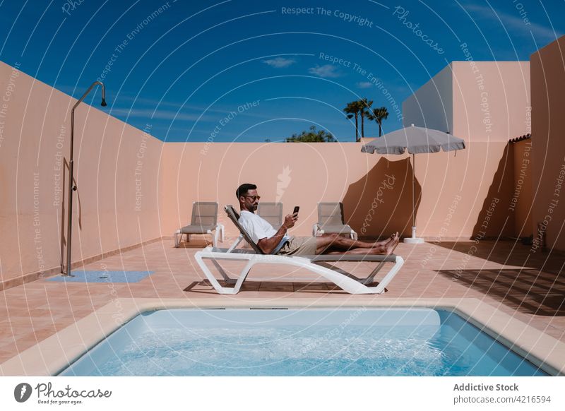 Mann liegt auf einem Liegestuhl und schaut auf seinem Smartphone am Pool herum freiberuflich Beckenrand Telearbeit Browsen Sommer männlich Surfen Mobile