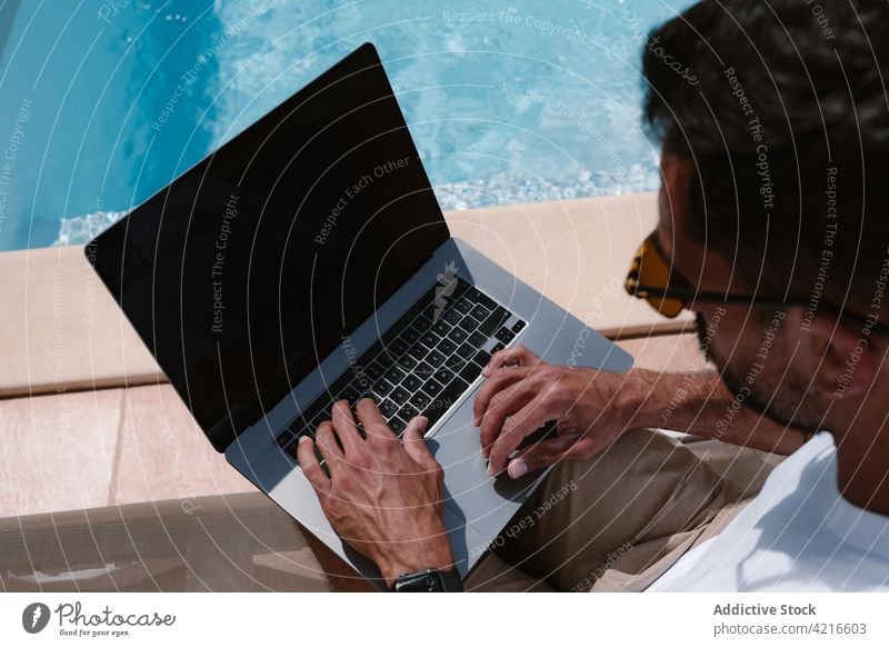 Mann liegt auf einem Liegestuhl und surft auf einem Computer am Pool freiberuflich Beckenrand Telearbeit Browsen Sommer männlich Internet online benutzend Lügen
