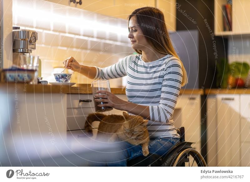 Behinderte junge Frau in der Küche mit Katze auf dem Schoß Rollstuhl häusliches Leben Behinderung deaktiviert Selbstvertrauen unabhängig im Innenbereich