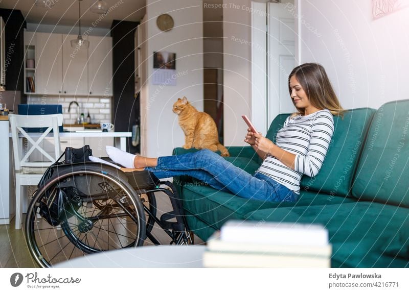 Behinderte junge Frau verwendet Smartphone, während sie zu Hause auf dem Sofa sitzt Rollstuhl häusliches Leben Behinderung deaktiviert Selbstvertrauen