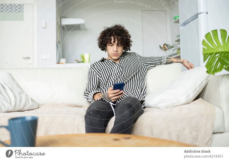 Tätowierter Mann surft zu Hause auf dem Sofa mit seinem Smartphone im Internet online Freizeit Surfen Tattoo heimwärts benutzend Apparatur Gerät zuschauend