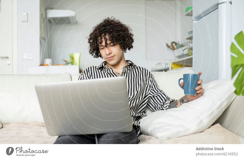 Tätowierter Mann surft im Internet auf einem Laptop auf dem Sofa zu Hause online Freizeit Surfen Tattoo heimwärts benutzend Apparatur Gerät zuschauend Kaffee